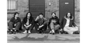 Black and white photo of the band Coco Jumbo. 5 members in the band. Sitting on the street smiling.