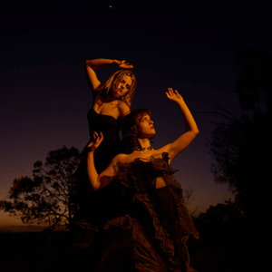 Press photo of Charm of Finches taken at dusk outside in the Australian landscape with gumtrees in the background. Ivy and Mabel from duo are dressed in fancy gowns while striking dramatic poses.