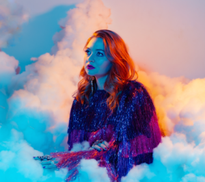 Audrey is shown from the waist up in a cloud. She is wearing a purple top and is lit up with blue light.