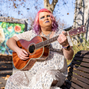 Image shows Joanna, aka Saint Ergo, with pink and purple hair and a white dress with floral print. She is sitting on a park bench playing her guitar.