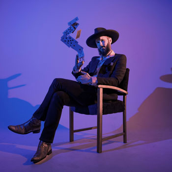 Press photo of Nigel Wearne. Purple background with Nigel sitting on a chair looking quite dapper dressed in a suit, cowboy boots and a wide flat brimmed hat while throwing a pack of cards.