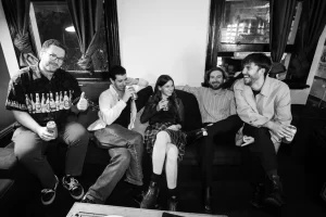 Black & white photo of the band The Slingers sitting on a couch laughing & drinking beer.