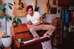 Gregor wears a red beret, white tee and beige pants and is sitting on a piano, feet on the keys, with guitars on the wall behind him.