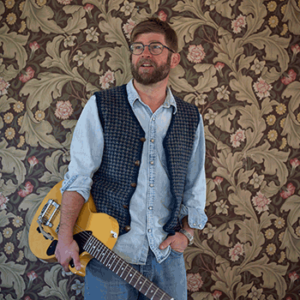 Colour photo of Old man Luedecke standing in front of a wall of floral brown wallpaper. He is holding a yellow guitar in his right arm and his left hand is in his pocket. Wearing blue denim shirt, blue woollen vest and blue jeans.