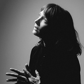 Black and white studio portrait of Sarah Blasko. Side profile with Sarah looking up towards the top left hand corner of the frame while gently rubbing her fingers together.