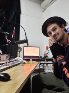 Timmus at the radio console, smiling and wearing a hat